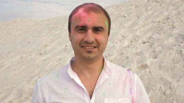 Porträtt av en glad ung man som blir beströdd med ljusa holi pulver av någon. Mot bakgrund av vit sand och blå himmel. — Stockvideo