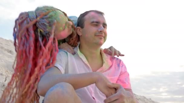 Портрет веселої молодої пари, що сидить на білому піску. Обличчя та одяг пофарбовані у яскраві кольори. Фестиваль Холі. — стокове відео