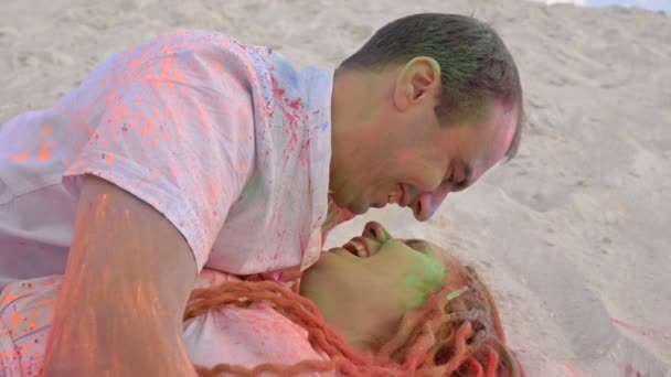 Jovem casal alegre encontra-se abraçando na areia. Faces e roupas são pintadas com pó multicolorido. Guy e menina olham um para o outro com amor e ternura. Holi Festival . — Vídeo de Stock