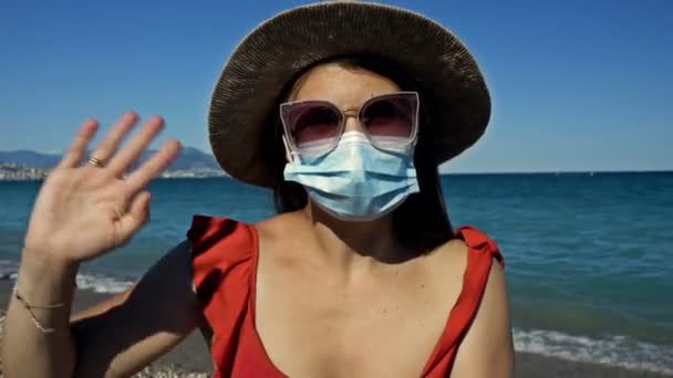 Młoda kobieta w okularach przeciwsłonecznych i masce ochronnej grzecznie macha rękami. Stoi na brzegu morza. Turystyka podczas koronawiru. — Wideo stockowe