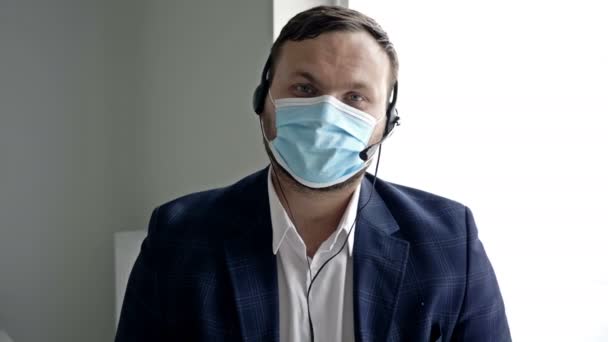 Man van middelbare leeftijd in een zakenpak, met een koptelefoon en een medisch masker. Online communicatie tijdens de coronavirusepidemie. — Stockvideo