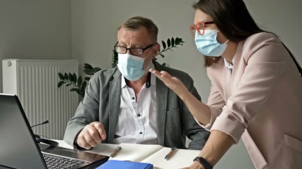 Hombre mayor discutiendo trabajo con su joven colega. Ambos con máscaras médicas. Trabajo de oficina durante la epidemia de covidio-19. — Vídeos de Stock