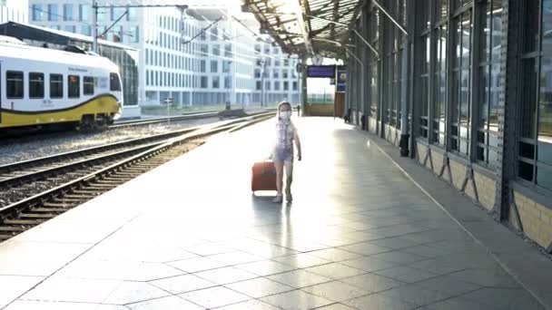 5-6 anos de idade menina está arrastando uma grande mala vermelha ao longo da plataforma ferroviária. A criança tem uma máscara médica no rosto. Viagens, turismo durante a epidemia de COVID-19. — Vídeo de Stock