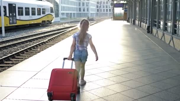 5-6 ετών κορίτσι σέρνει μια μεγάλη κόκκινη βαλίτσα κατά μήκος της σιδηροδρομικής πλατφόρμας. Πίσω όψη. — Αρχείο Βίντεο
