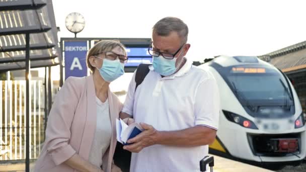 Starší manželský pár v lékařských maskách na tvářích stojí na nástupišti a čeká na vlak. Cestování, cestovní ruch během epidemie COVID-19. — Stock video