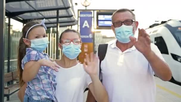 Eine dreiköpfige Familie mit Schutzmasken im Gesicht trifft am Bahnhof auf jemanden. Eine junge Frau, ihr Vater und ihre kleine Tochter winken glücklich mit den Händen. — Stockvideo