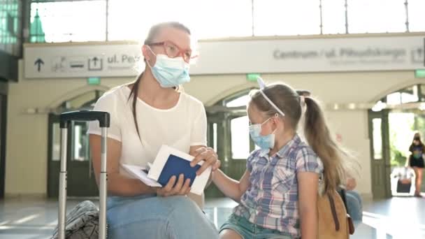 Молода жінка з маленькою донькою чекає свого польоту в кімнаті очікування. Обидва мають медичну маску на обличчі. Подорожі, туризм під час епідемії коронавірусу . — стокове відео
