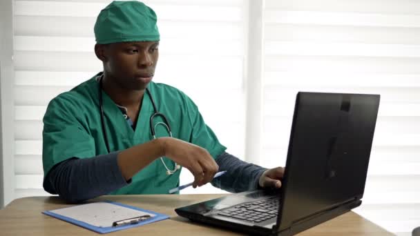Jonge zwarte arts of stagiair werkt op een computer en maakt aantekeningen in zijn kantoor. — Stockvideo