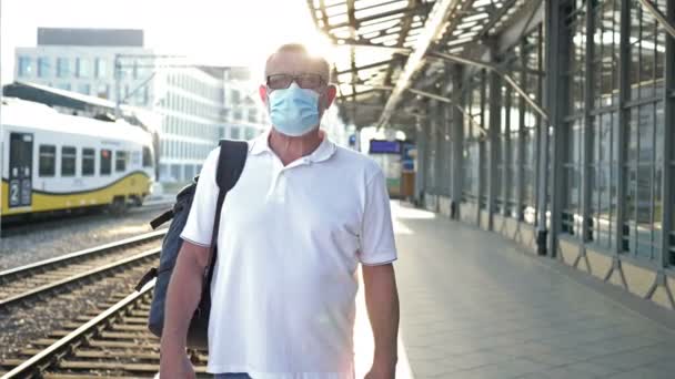 彼の顔に医療マスクをした高齢者の乗客。彼は列車を待っているホームに立っている。旅行、 COVID-19流行中の観光. — ストック動画