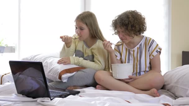 Two girlfriends of teenage age watching dramatic movie at home, sitting on the bed and eating ice cream. They are both moved and cry. — Stock Video