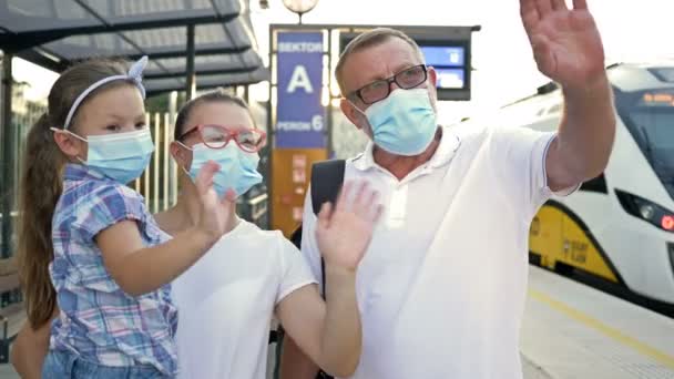 Familie van drie, met beschermende maskers op hun gezicht, ontmoet iemand op het station. Een jonge vrouw, haar vader en dochtertje zwaaien gelukkig met hun handen. — Stockvideo