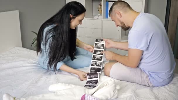 Manželé sedí v ložnici na posteli a dívají se na ultrazvuk nenarozeného dítěte. — Stock video