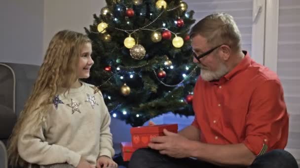 Nagyapa és unokája a karácsonyfa mellett. A lány kinyitja az ajándékot, és örül. Boldog új évet! — Stock videók