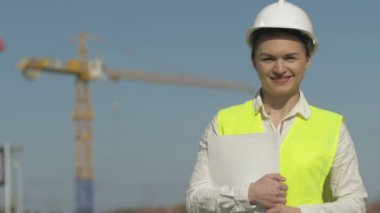 İnşaat alanında koruyucu giysiler giyen kız müteahhit. Kızın elinde belgeler ve gülücüklerle dolu bir klasör var.