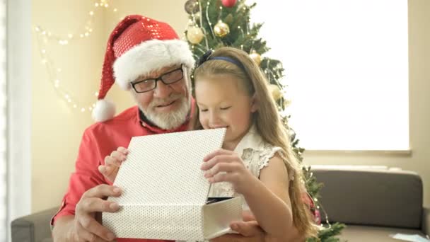 Дедушка в шляпе Санты дарит рождественский подарок своей маленькой внучке. Ребенок счастлив. На фоне рождественской елки. Счастливого Рождества. С Новым годом. — стоковое видео