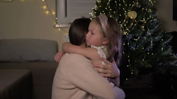 Mam knuffelt een huilend dochtertje tegen de achtergrond van een elegante kerstboom. De baby is ergens van streek door.. — Stockvideo