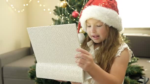 Glückliches kleines Mädchen mit Weihnachtsmütze auf dem Hintergrund des Weihnachtsbaums. Das Kind freut sich über das Weihnachtsgeschenk. — Stockvideo