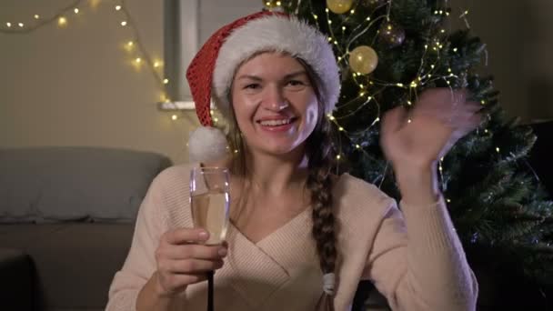 Beautiful cheerful woman in santa hat with a glass of champagne. Against the background of a Christmas tree. Happy New Year and Merry Christmas. — Stock Video