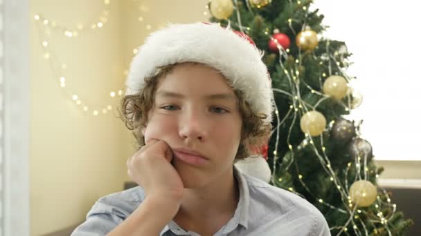 Egy Mikulás sapkás tinédzser fiú portréja, aki unatkozik vagy szomorú. A karácsonyfa hátterében. Boldog karácsonyt! Boldog új évet!. — Stock videók