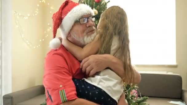 A menina abraça o avô em Santa Hat. Contra o fundo da árvore de Natal. — Vídeo de Stock