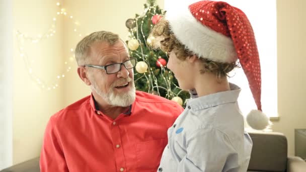 Підліток, який носить капелюха Санти, обіймає свого діда. На тлі ялинки. Веселого Різдва. З Новим роком. — стокове відео
