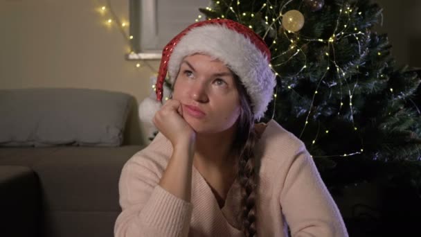 美しいです中世の女性で赤いパーティーサンタ帽子座っていますザクリスマスツリーの近くに悲しい表情で顔. — ストック動画