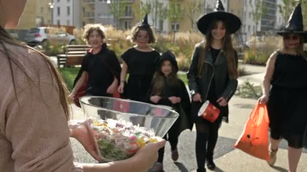 Vrouw trakteert snoep op een groep kinderen in zwarte mooie jurk met enge make-up op hun gezicht. Halloween tradities. — Stockvideo