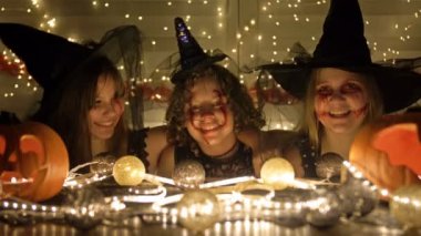 Yüzlerinde korkunç makyajlar olan üç siyah cadı kostümlü genç kız bir masada oturup şarkılar söylüyorlar. Masadaki Cadılar Bayramı özellikleri.