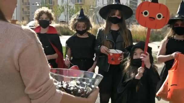 Eine Frau verteilt Süßigkeiten an eine Gruppe Kinder in schwarzen Verkleidungen mit gruseligem Make-up im Gesicht. Halloween-Traditionen. — Stockvideo