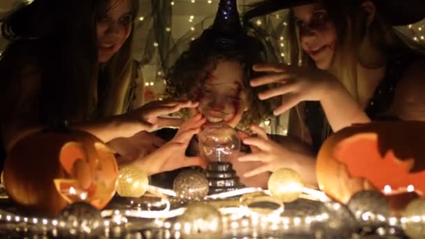 Three teenage girls dressed as witches with scary makeup on their faces are doing magic. Dark background. Halloween attributes on the table. — Stock Video