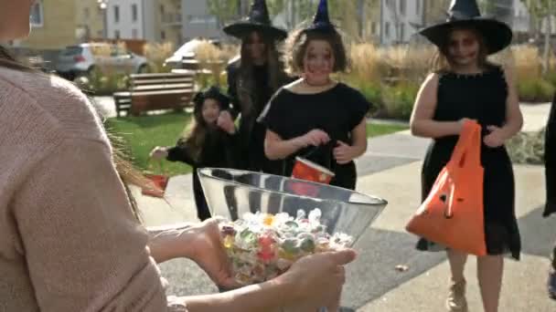 女性は顔に怖いメイクの黒い空想の服を着た子供たちのグループにお菓子を扱います。ハロウィンの伝統. — ストック動画