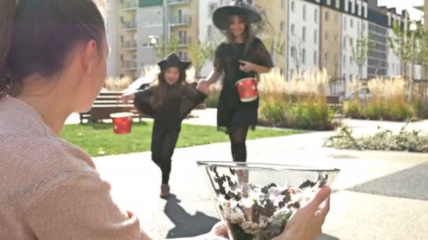 女は黒の派手なドレスで2人の姉妹にお菓子を扱う。子供たちは喜んでお菓子でバケツを満たします。ハロウィンの伝統. — ストック動画