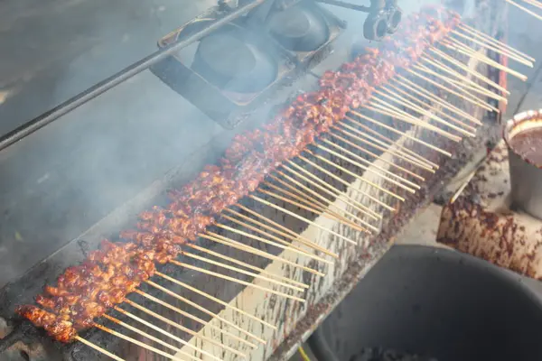 Satay with goat meat prepared on a charcoal grill. Smoke billows from the grill.
