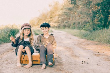 İki çocuk kırsal bir yolda eğleniyor. Bir kız ve bir erkek büyük sarı bavula oturuyor. Retro tarzı
