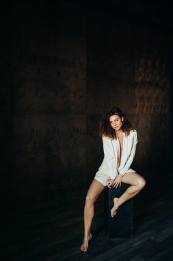 Güzel bir genç kız iç çamaşırı ve karanlık bir stüdyoda bir ceket portresi. Kıvırcık saçlı. Düğmeleri gömlek