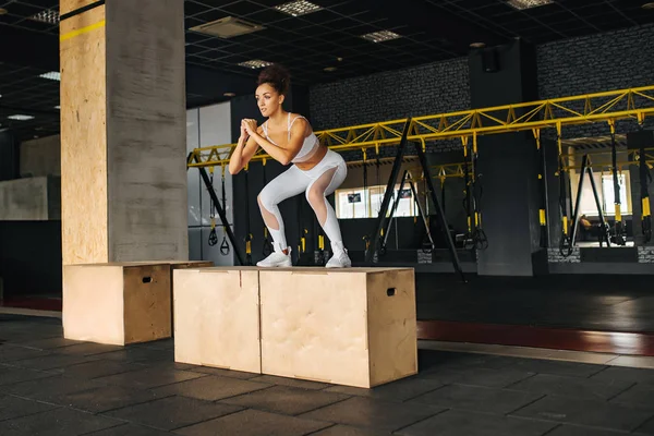 Cross fit training. Sporty fitness girl in black sportswear with perfect body jumping over some boxes in fitness gym. Woman jumping at crossfit style club. Female athlete is performing jumps