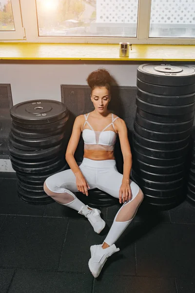 Fitness girl sitting between heavy weights for the barbell. Sport fashion model. Proper nutrition. Sport fitness workout. Fitness woman in white sportswear top and leggins
