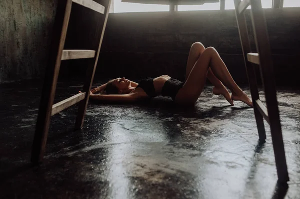 Dark room portrait of perfect young woman in sexy black lingerie. Woman with long charming legs lying in loft room