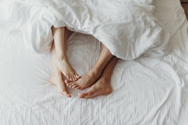 Yatakta çıplak ayakla yatıp battaniyenin altına dokunan bir çift. Yakınlığın, sevginin ve sabah sakinliğinin sembolü. Yumuşak ışık yumuşak ve rahat bir ruh halini vurgular.