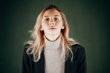 Balıkçı yaka kazak ve ceket giyen genç bir kadın koyu yeşil arka planda poz veriyor. Doğal ifadeyle özgüvenli ve şık bir portre..