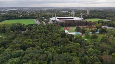 Uçsuz bucaksız bir şehir parkı ve ormanın gökyüzü. Ufukta ağaçlarla çevrili bir stadyum, atletizm pisti ve havuzları var. İnsansız hava aracı manzarası, gün ışığı, insan yok..