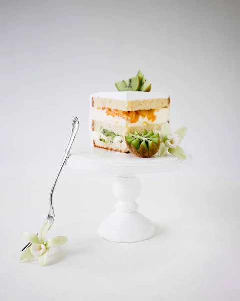 Piece of biscuit cake with tropical fruits kiwi and peaches on white wooden cake stand