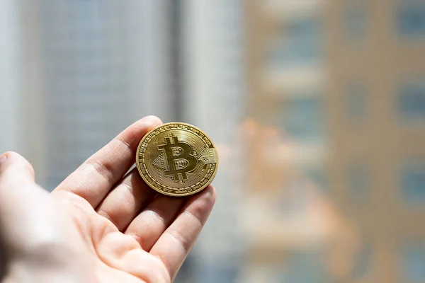 Bulanık şehir arkaplanına karşı elinde altın Bitcoin para tutan kişinin yakın plan fotoğrafı, dijital finans ve gelecekteki parayı simgeliyor. 
