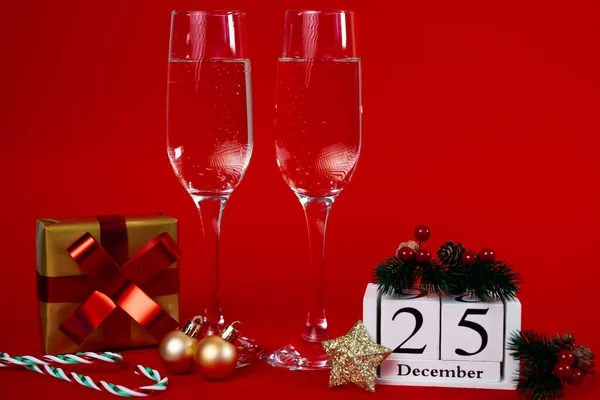 Festive red holiday arrangement featuring champagne flutes, ornaments, gift and block calendar showing 25. Christmas countdown concept. 