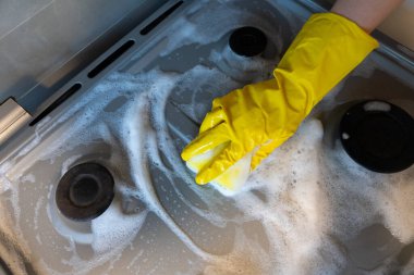 Hand in protective gloves scrubbing gas stove with sponge and soap foam, household cleaning and hygiene concept. 