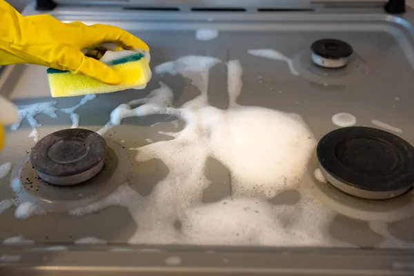 Close-up of gloved hand scrubbing dirty gas stove burner with sponge and detergent foam, concept of kitchen hygiene and disinfection.