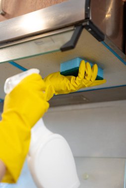 Woman in yellow gloves cleaning extractor hood filter in kitchen. Hygiene, household maintenance, and safety concept. 