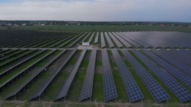 Αεροφωτογραφία drone σε μεγάλους ηλιακούς συλλέκτες σε ένα ηλιακό αγρόκτημα σε συννεφιασμένο καλοκαιρινό βράδυ. Ηλιακοί σταθμοί ηλεκτροπαραγωγής, πράσινο γρασίδι. Βίντεο βίντεο 4k. — Αρχείο Βίντεο