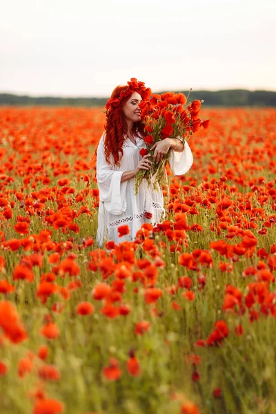 Portrait of beautiful redhead woman in wreath with bouquet of poppies on green field in summer sunset