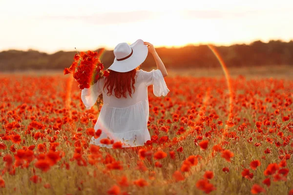 Redhead beautiful woman in hat with bouquet of poppies on green field in summer sunset